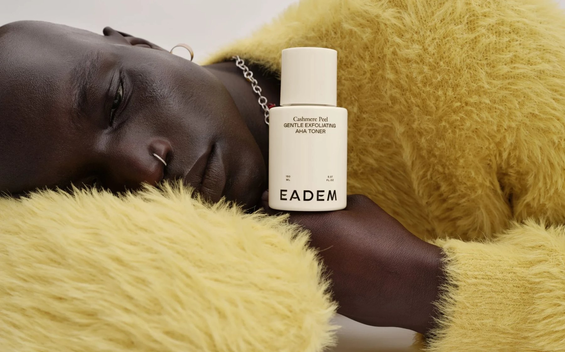 Woman in yellow sweater holds Eadem bottle to cheek.