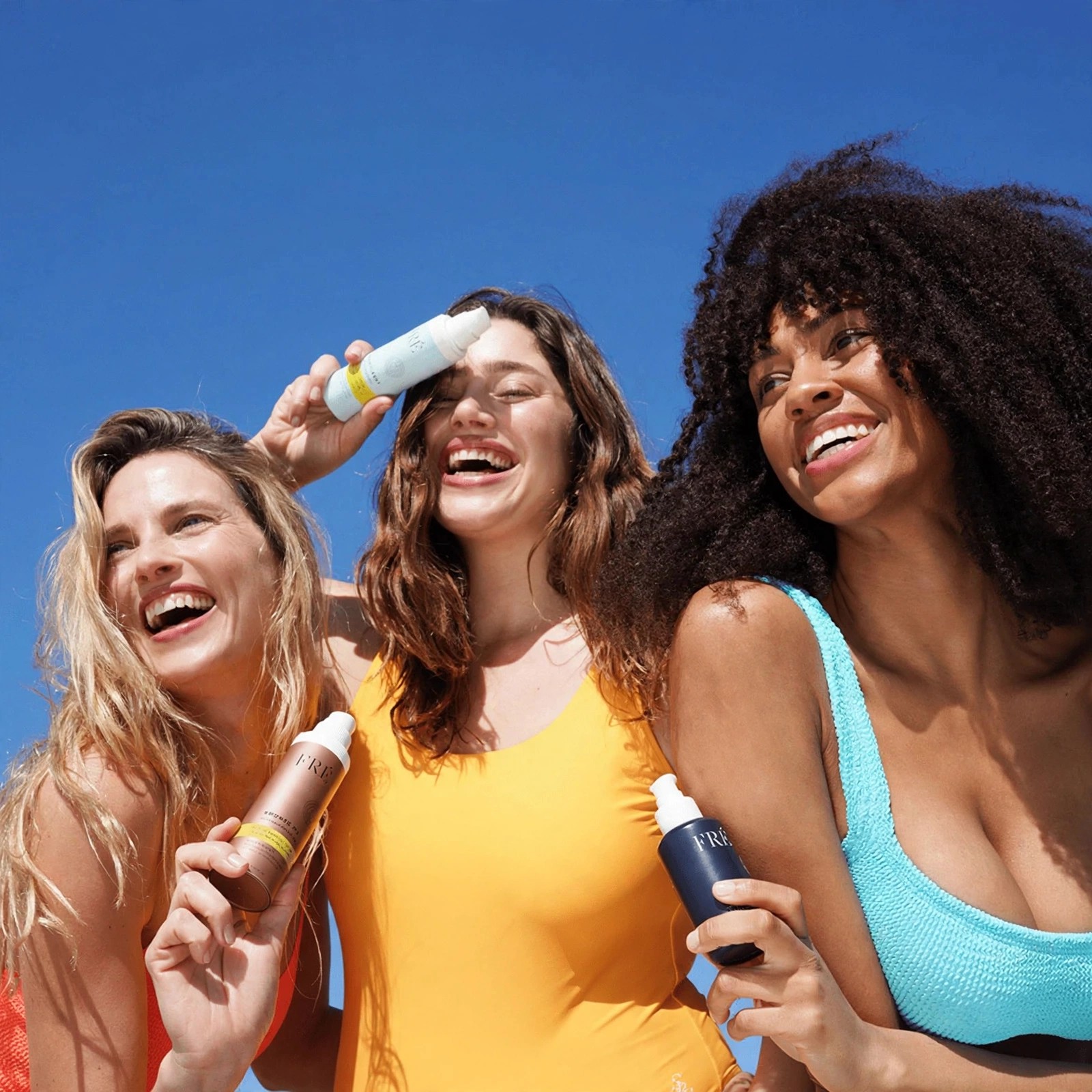 Three smiling women holding Fre products under blue sky.