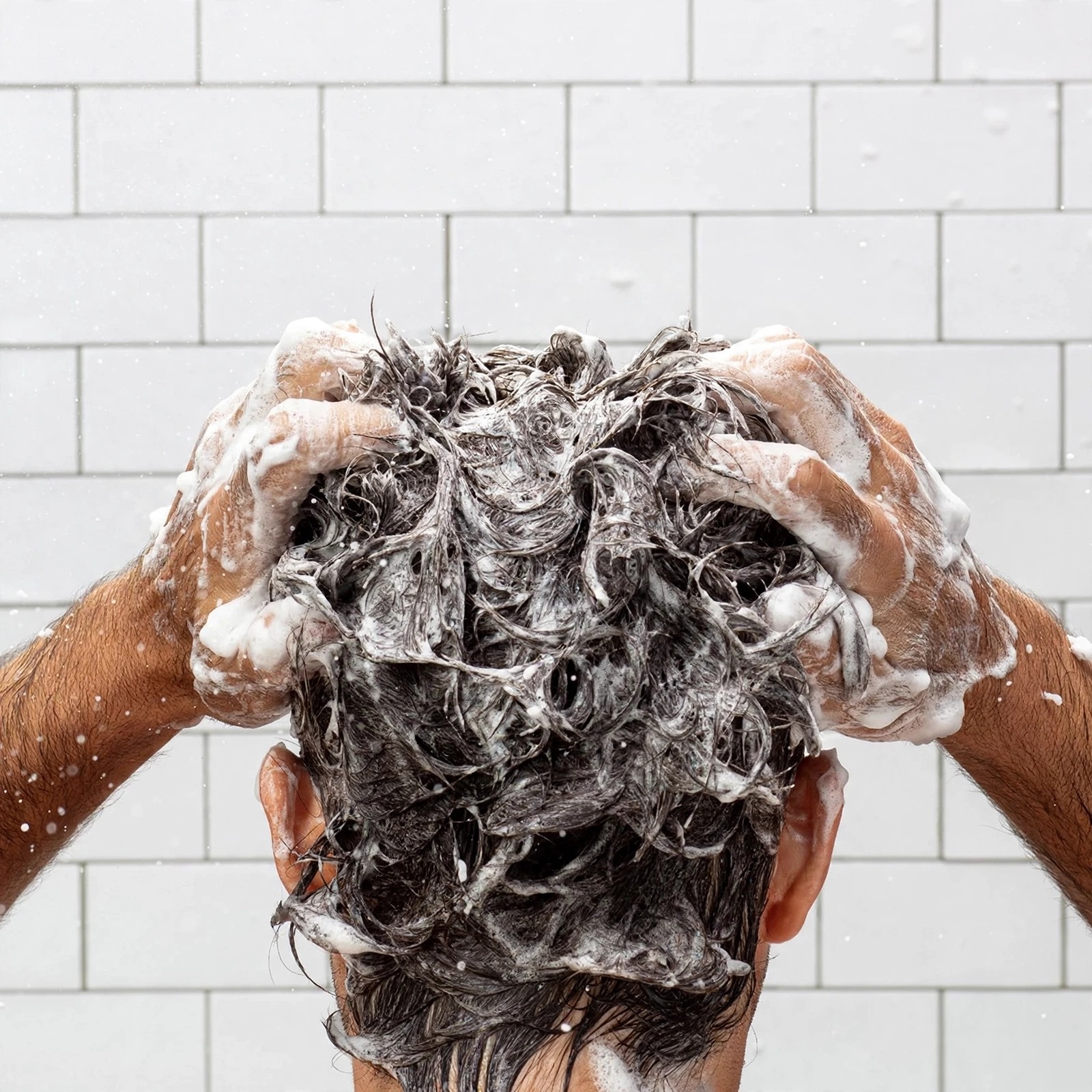 Hands lathering foamy product into wet hair in shower.