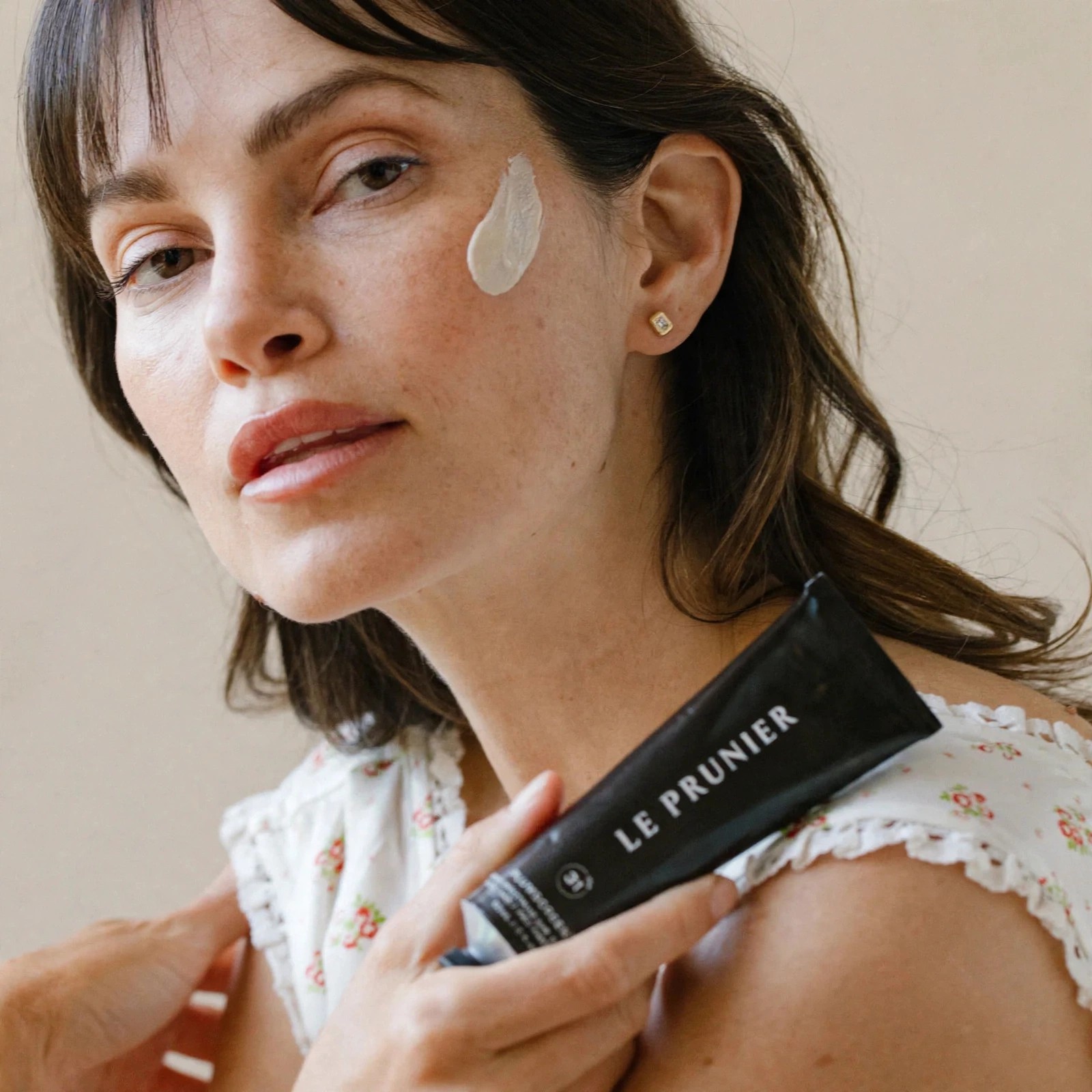 Woman holding Plumscreen tube with cream on cheek.