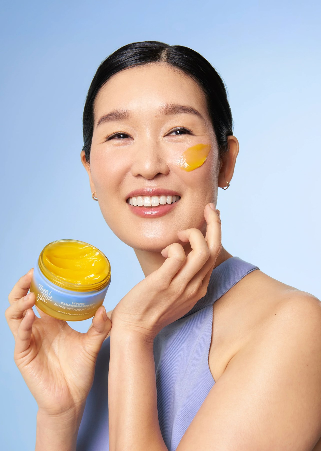 Woman holds cleansing balm jar with balm swatched on cheek.