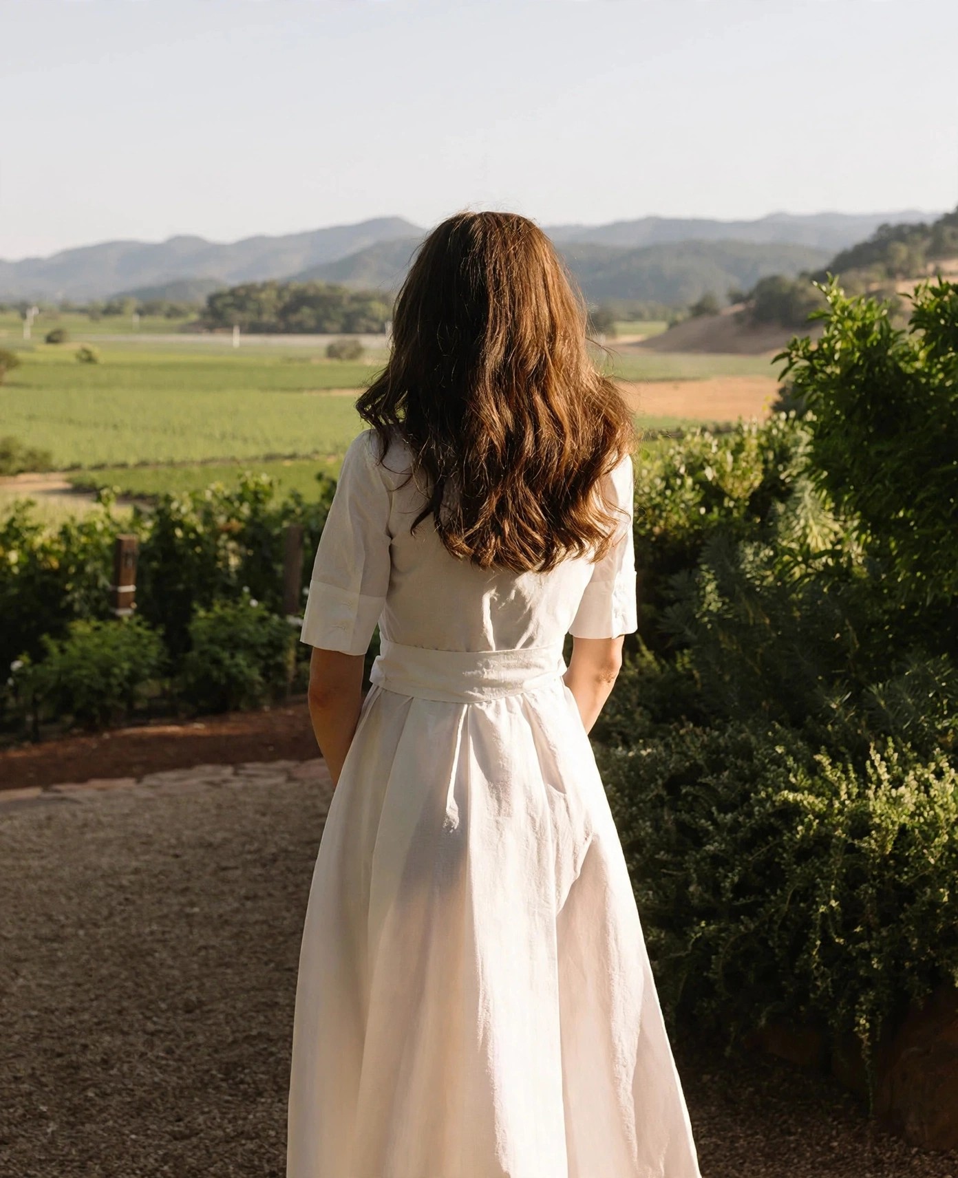 Woman in white dress overlooking vineyards and rolling hills.